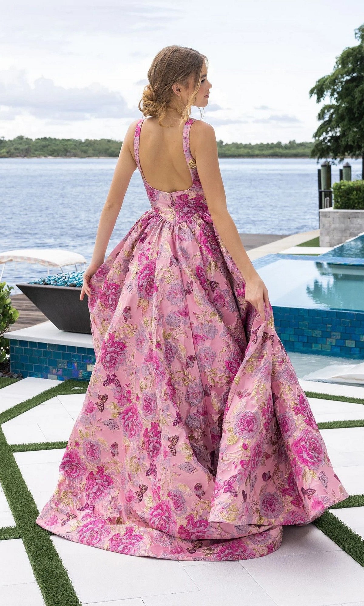 A woman wearing the Portia and Scarlett XO188 long formal dress in pink floral stands by a pool, holding her gown, with shimmering water and lush trees behind her.
